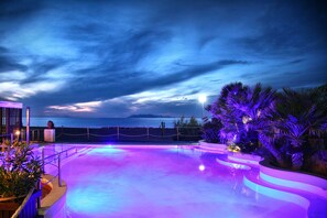 A swimming pool with a curved edge, surrounded by palm trees and a view of the ocean at dusk. - Hotel Il Gabbiano Beach (Terme Vigliatore)