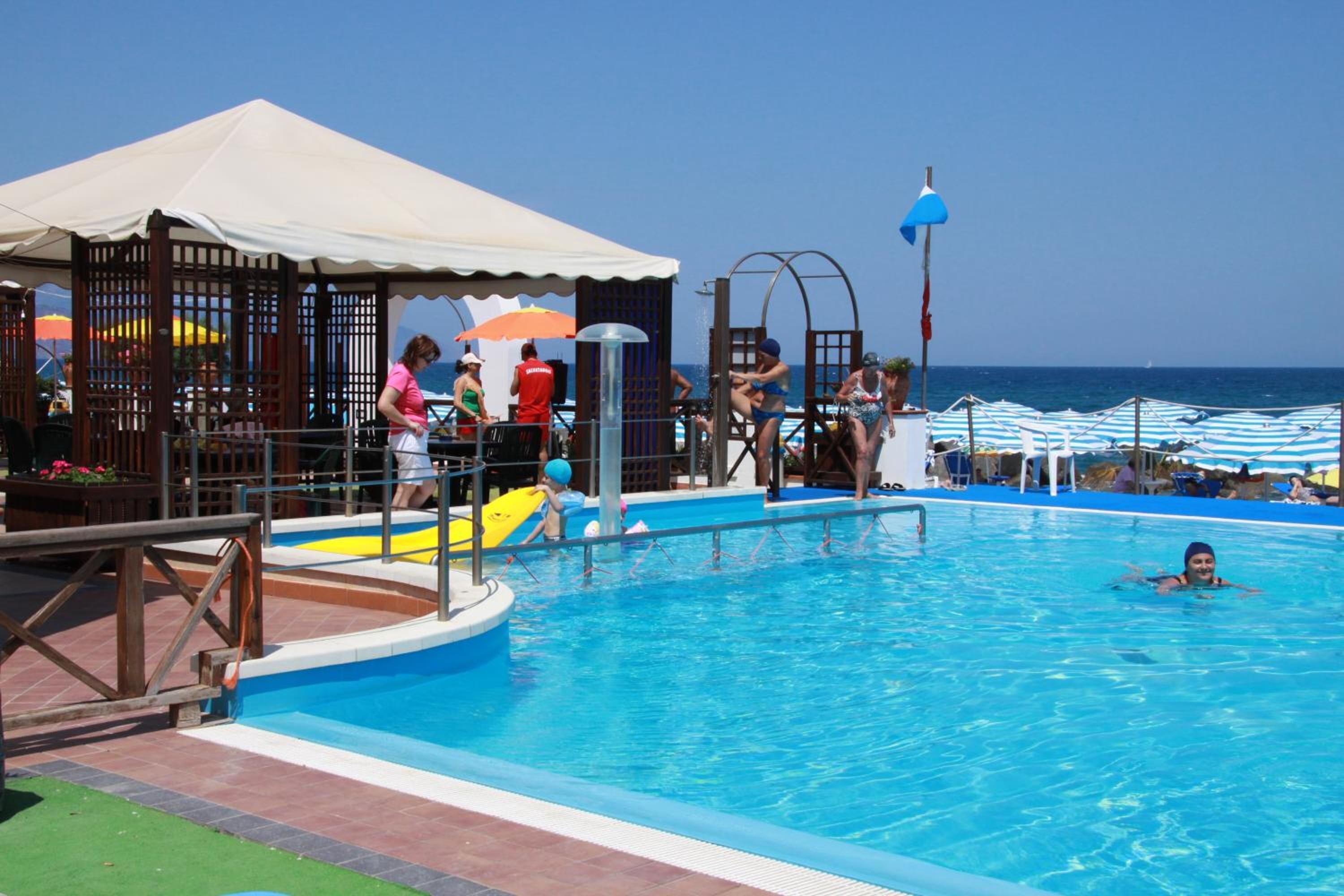 Zona de piscina amb un trampolí, una zona de seients coberta i vistes al mar.