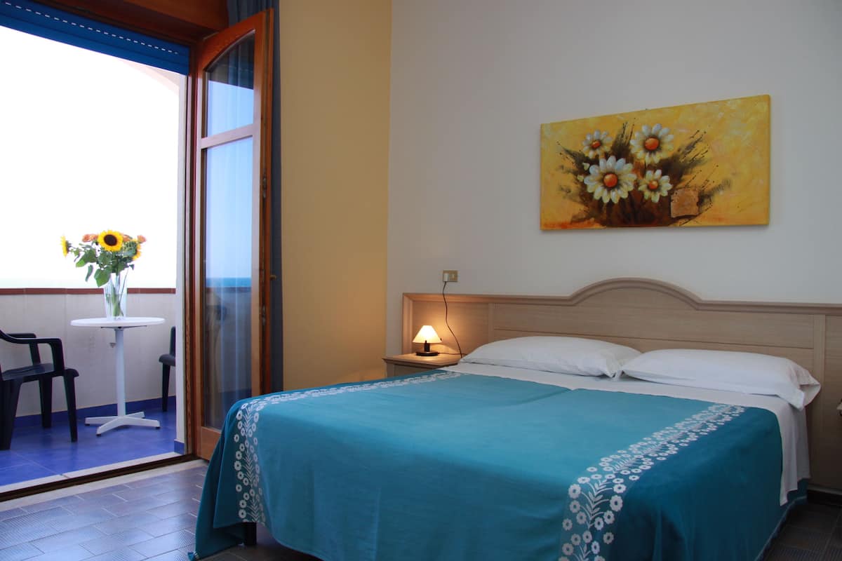 Ein Hotelzimmer mit Bett, Nachttisch mit Lampe, einem Blumenbild an der Wand, einem Fenster mit Aussicht und einem kleinen Tisch mit Stühlen draußen.
