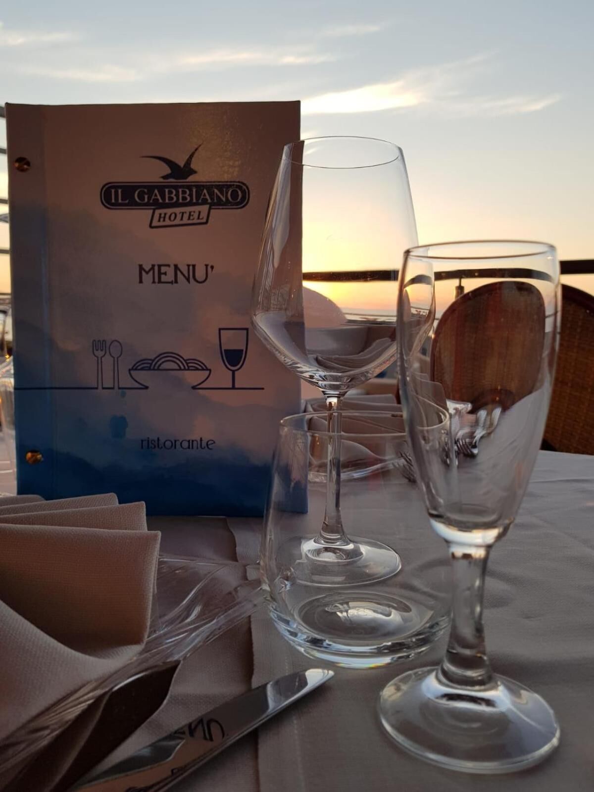 A table set with wine glasses and cutlery, a menu board for Il Gabbiano Hotel, and a view of the sunset.