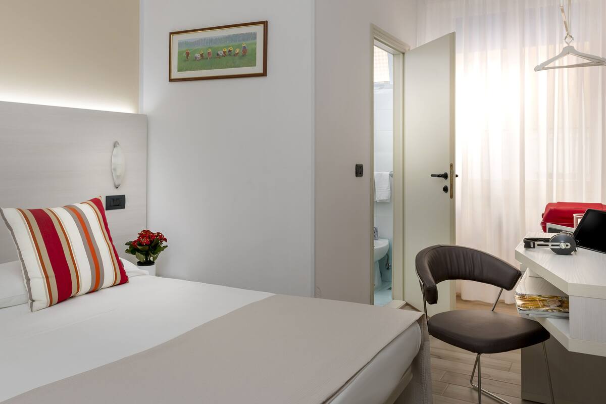 A hotel room with a bed, a desk, a chair, a painting on the wall, and a door leading to a bathroom.