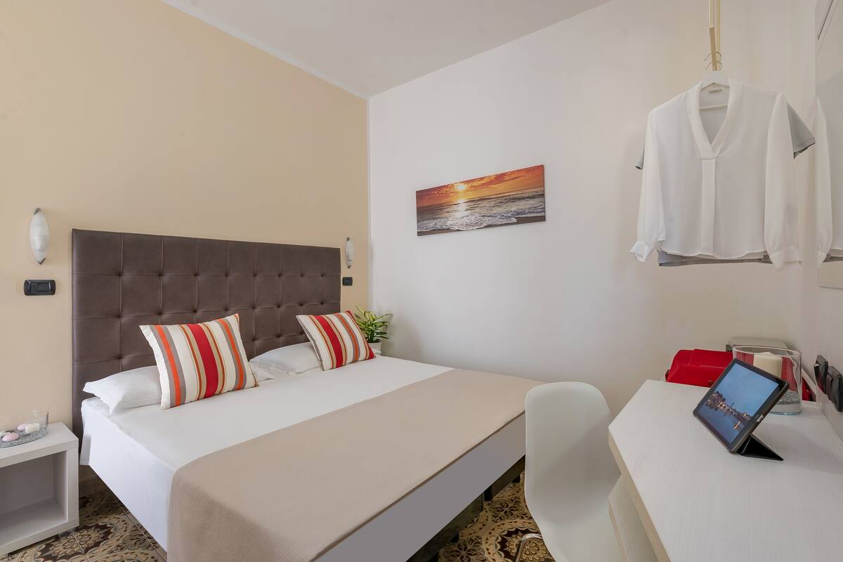 A hotel room with a bed, a bedside table, a desk with a tablet, a hanging robe, and a painting of a sunset.