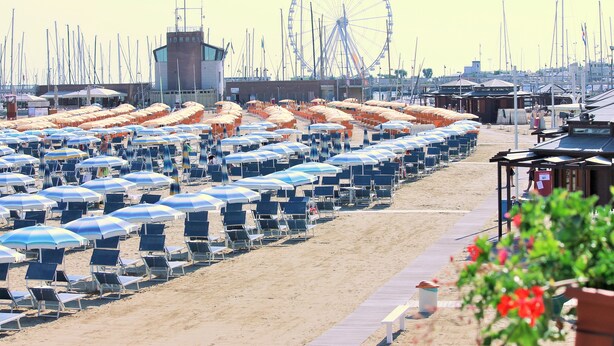 A beach with rows of blue and white striped umbrellas and beach chairs, a Ferris wheel, and a boardwalk.