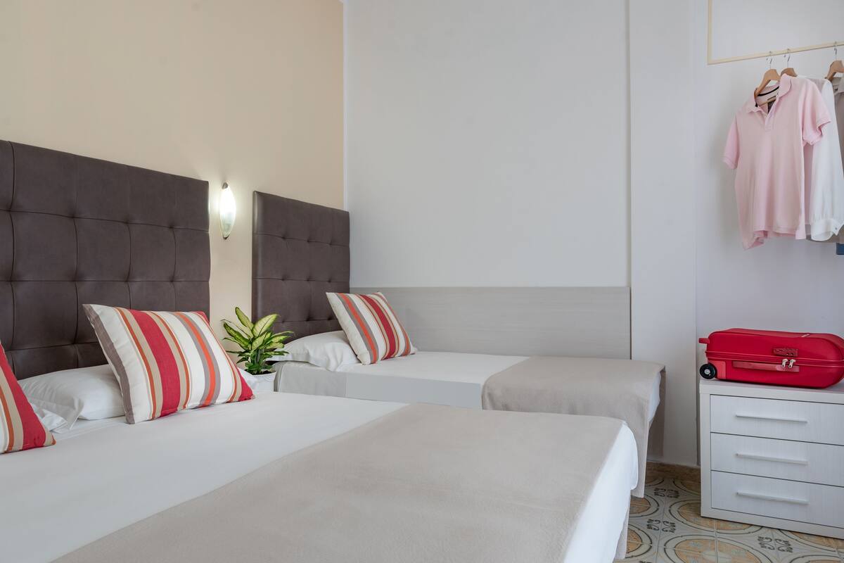 A hotel room with two beds, a grey headboard, striped pillows, a small plant, and a red suitcase on a white dresser.