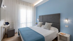 A modern bedroom with a large bed, a bedside table, a small desk with a laptop, and a window with sheer curtains.