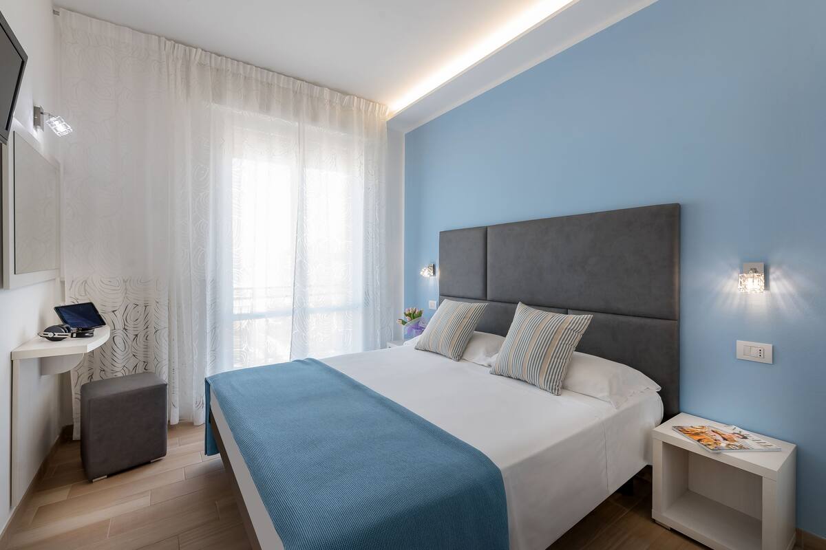 A modern bedroom with a large bed, a bedside table, a small desk with a laptop, and a window with sheer curtains.
