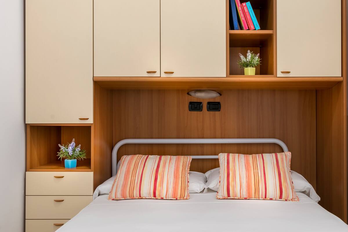 A bedroom with a bed, two pillows, a nightstand, a shelf with books and a plant, and a wardrobe.
