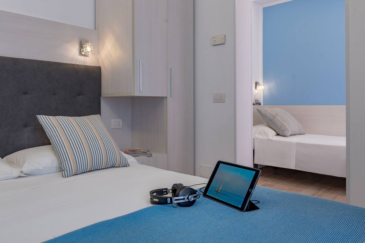 A modern bedroom with a bed, a desk with a laptop, and headphones.