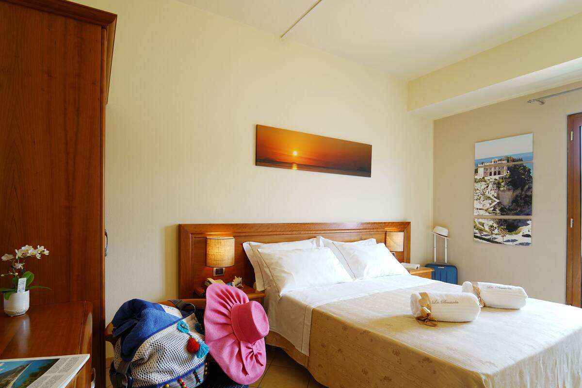 Ein Hotelzimmer mit einem Bett, einem Nachttisch, einer Lampe, einem Koffer, einem rosa Hut und einem Gemälde vom Sonnenuntergang.