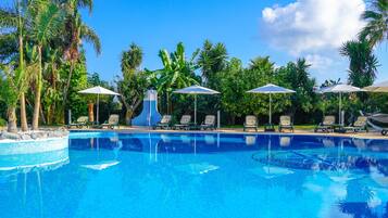 Una piscina azul cristalina con sillas de descanso y sombrillas, rodeada de palmeras y vegetación.