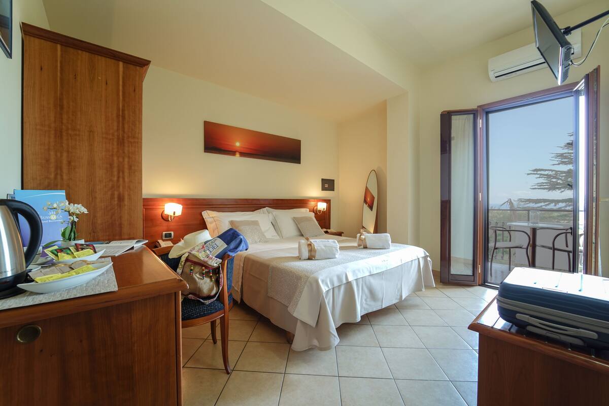 Ein Hotelzimmer mit Bett, Schreibtisch mit Wasserkocher, Stuhl, Tisch mit Blick und einer Schiebetür zum Außenbereich.