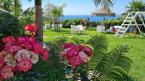 Un jardín exuberante con palmeras, una estructura de juegos blanca y vistas al océano.