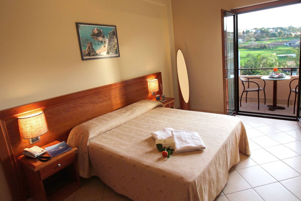 Ein Hotelzimmer mit einem großen Bett, einem Nachttisch mit Lampe, einem Beistelltisch mit Telefon, einem gerahmten Bild an der Wand und einem Balkon mit Tisch und Stühlen.