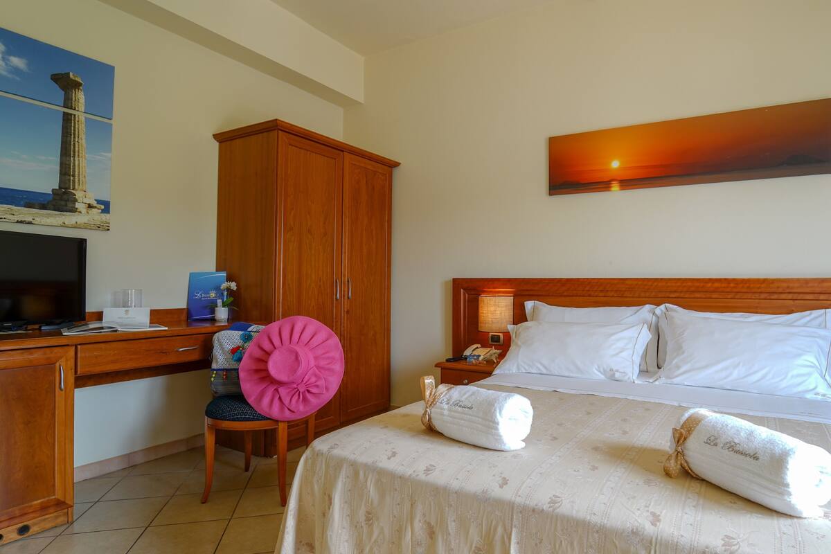 Ein Hotelzimmer mit Bett, Schreibtisch mit Computer, Stuhl, Kleiderschrank und einem Sonnenuntergangsbild.