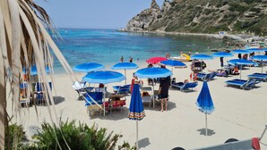 Una playa con sombrillas azules, sillas de descanso y un promontorio rocoso al fondo.