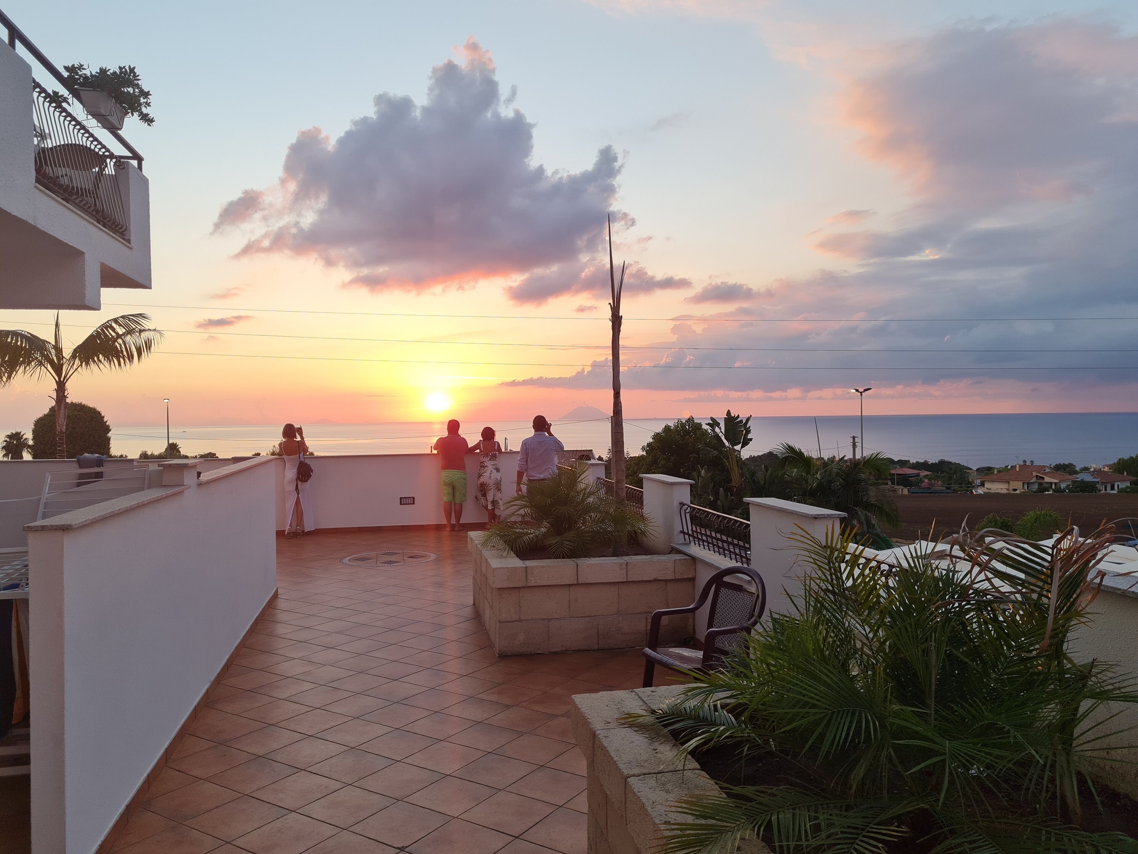 Una terraza con vista al atardecer sobre el mar, suelo de baldosas y algunas plantas en macetas.