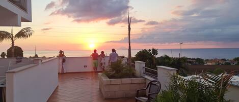 Una terraza con vista al atardecer sobre el mar, suelo de baldosas y algunas plantas en macetas.