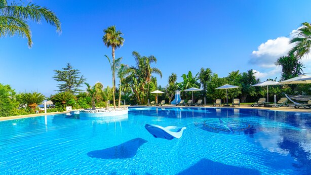 Una piscina azul cristalina con sillas de descanso y sombrillas, rodeada de palmeras y vegetación.