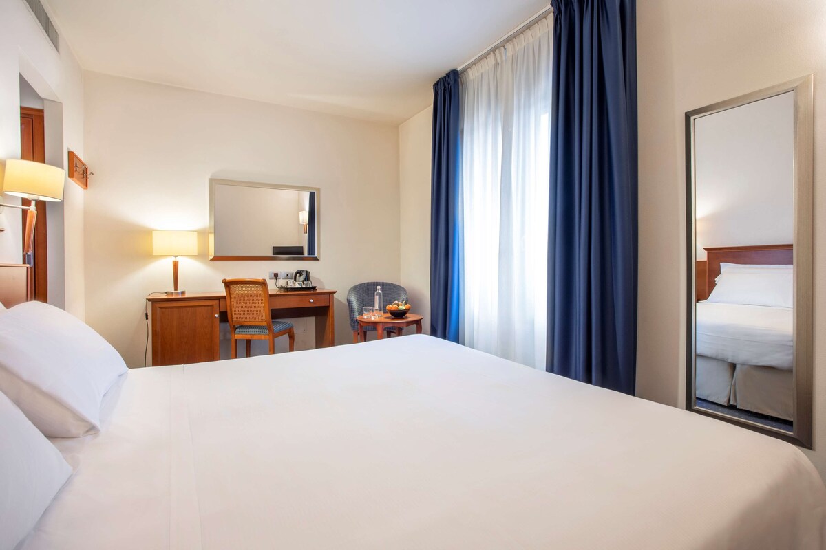 A hotel room with a large bed, a desk with a chair, a small table with a bowl of fruit, a lamp, and a mirror.