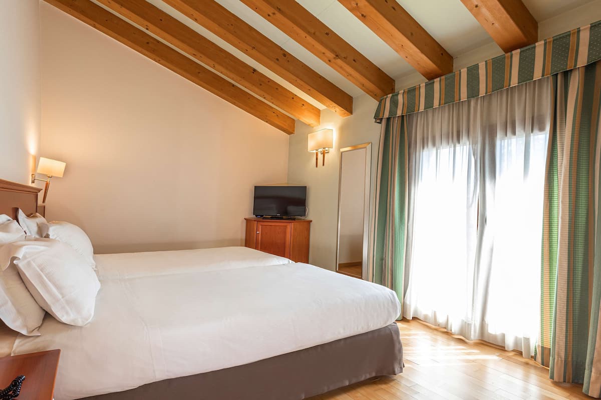 A hotel room with a large bed, wooden ceiling beams, a television on a wooden cabinet, and a window with striped curtains.