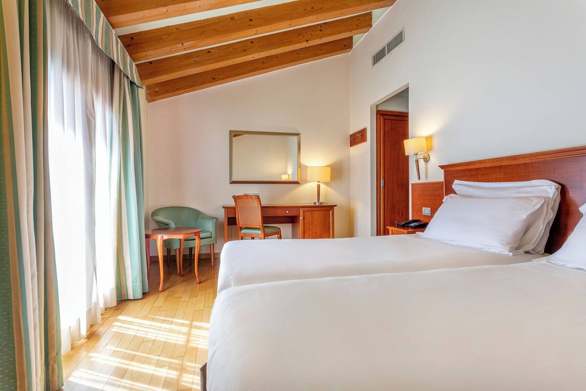 A hotel room with two beds, a desk, a chair, and a lamp.
