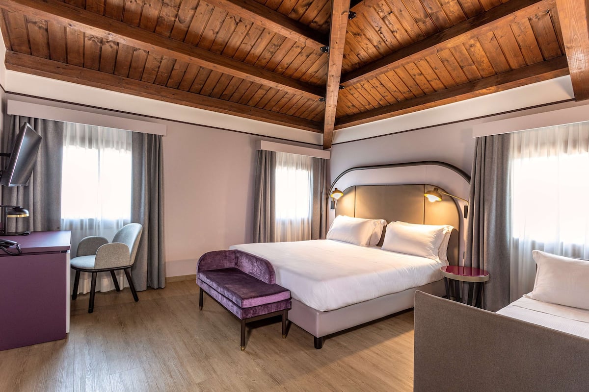 A hotel room with a wooden ceiling, a bed with white linens, a desk with a chair, and a small table with a lamp.