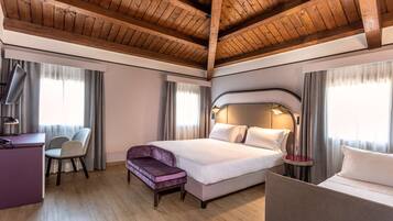 Habitación de hotel con techo de madera, cama con ropa blanca, escritorio con silla y una mesita con lámpara.