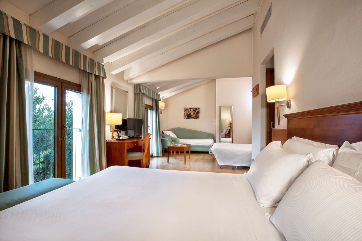 A hotel room with a large bed, a desk with a chair, a sofa, and a balcony with curtains.