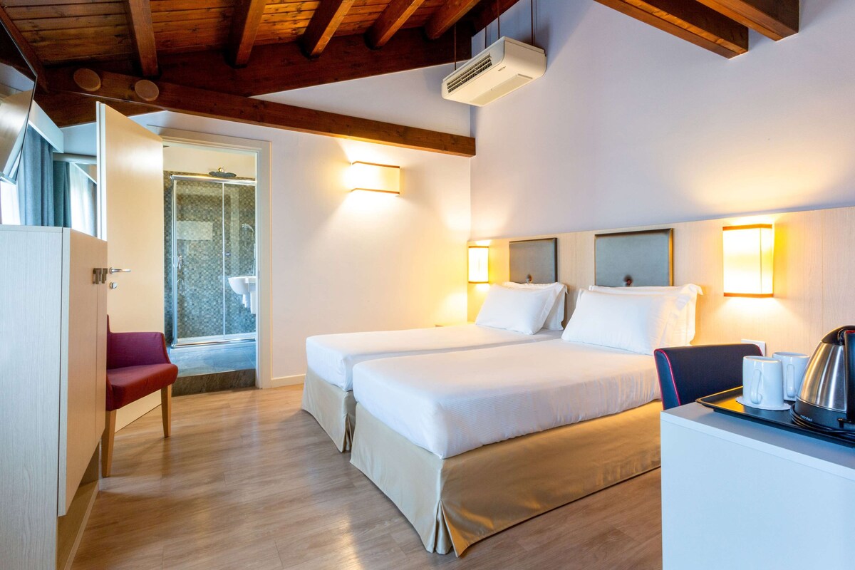 A hotel room with two beds, a desk with a kettle, and a shower area.