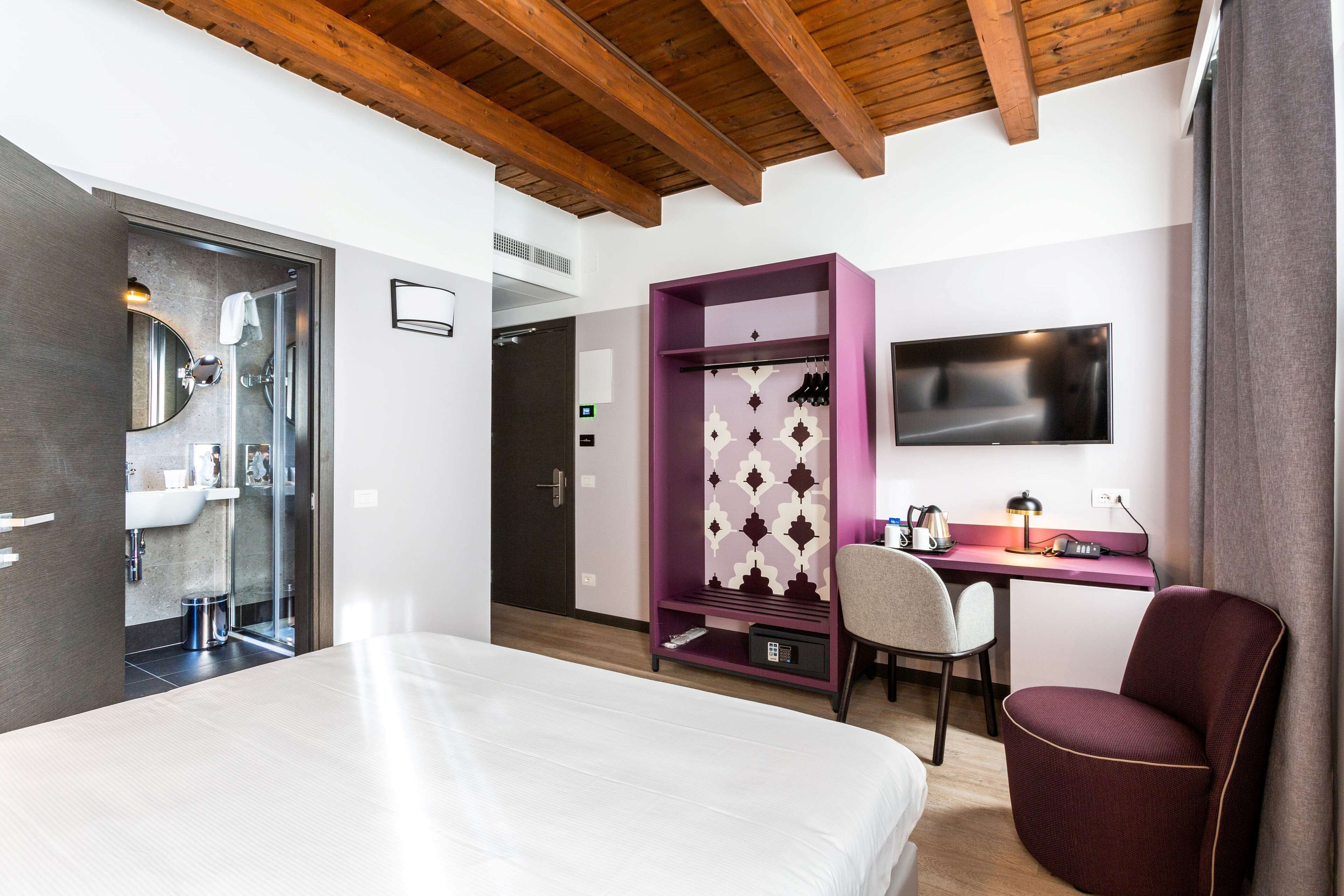 A modern hotel room with a bed, a desk with a TV, a purple wardrobe, and a shower area.