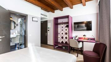 A modern hotel room with a bed, a desk with a TV, a purple wardrobe, and a shower area.