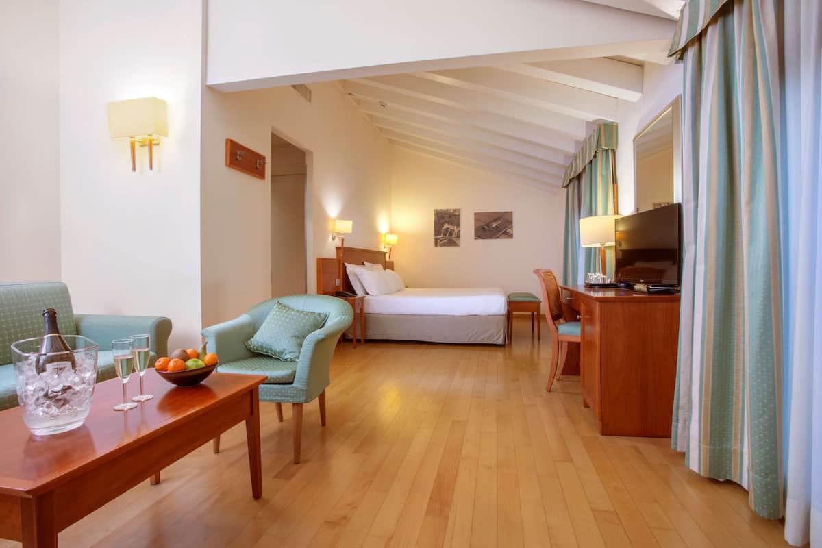 A hotel room with a wooden floor, a bed, a desk with a chair, a small table with a bowl of fruit and glasses, and a television.