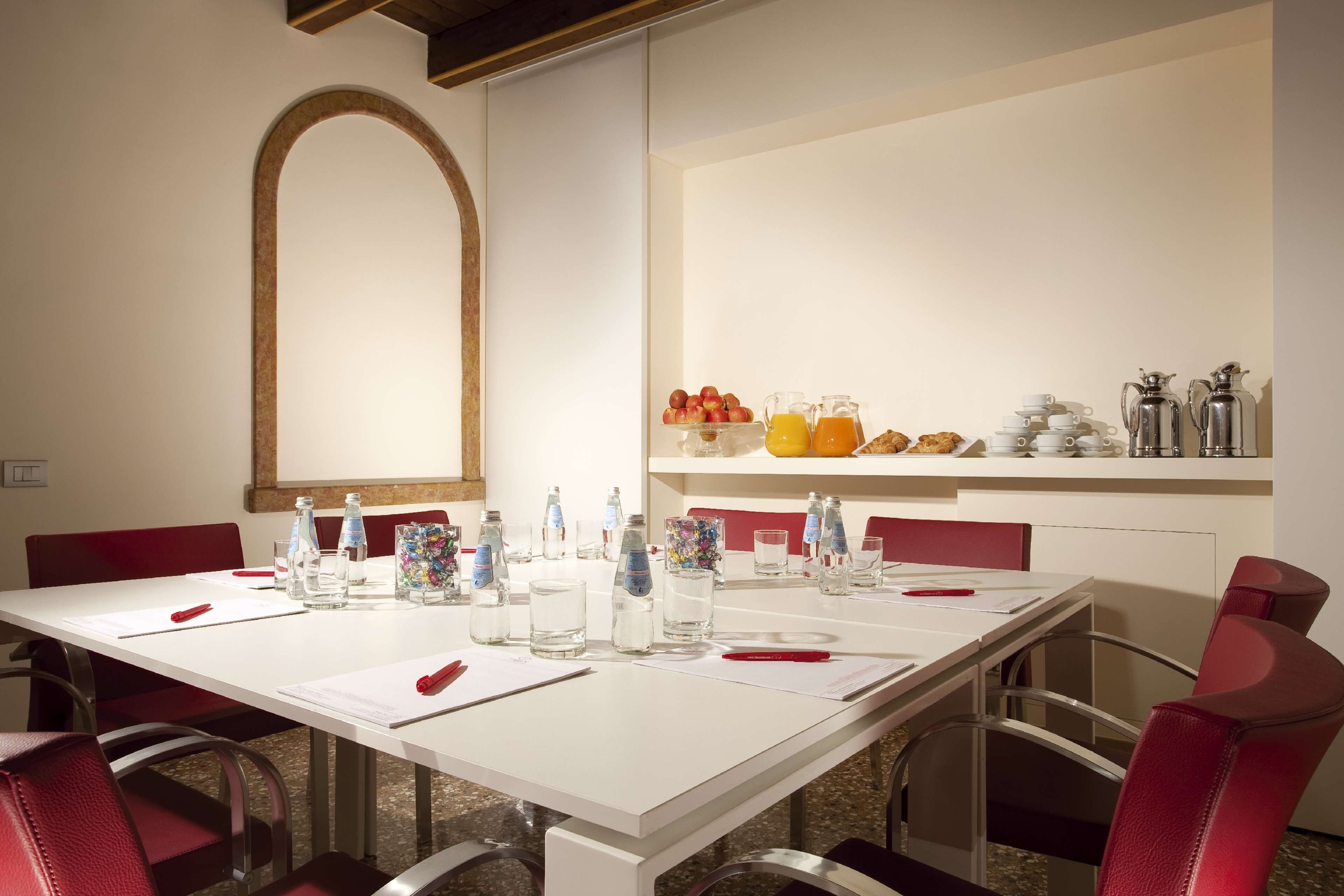 A conference room with a long table, red chairs, and a whiteboard. There are water bottles, glasses, and a fruit basket on the table.