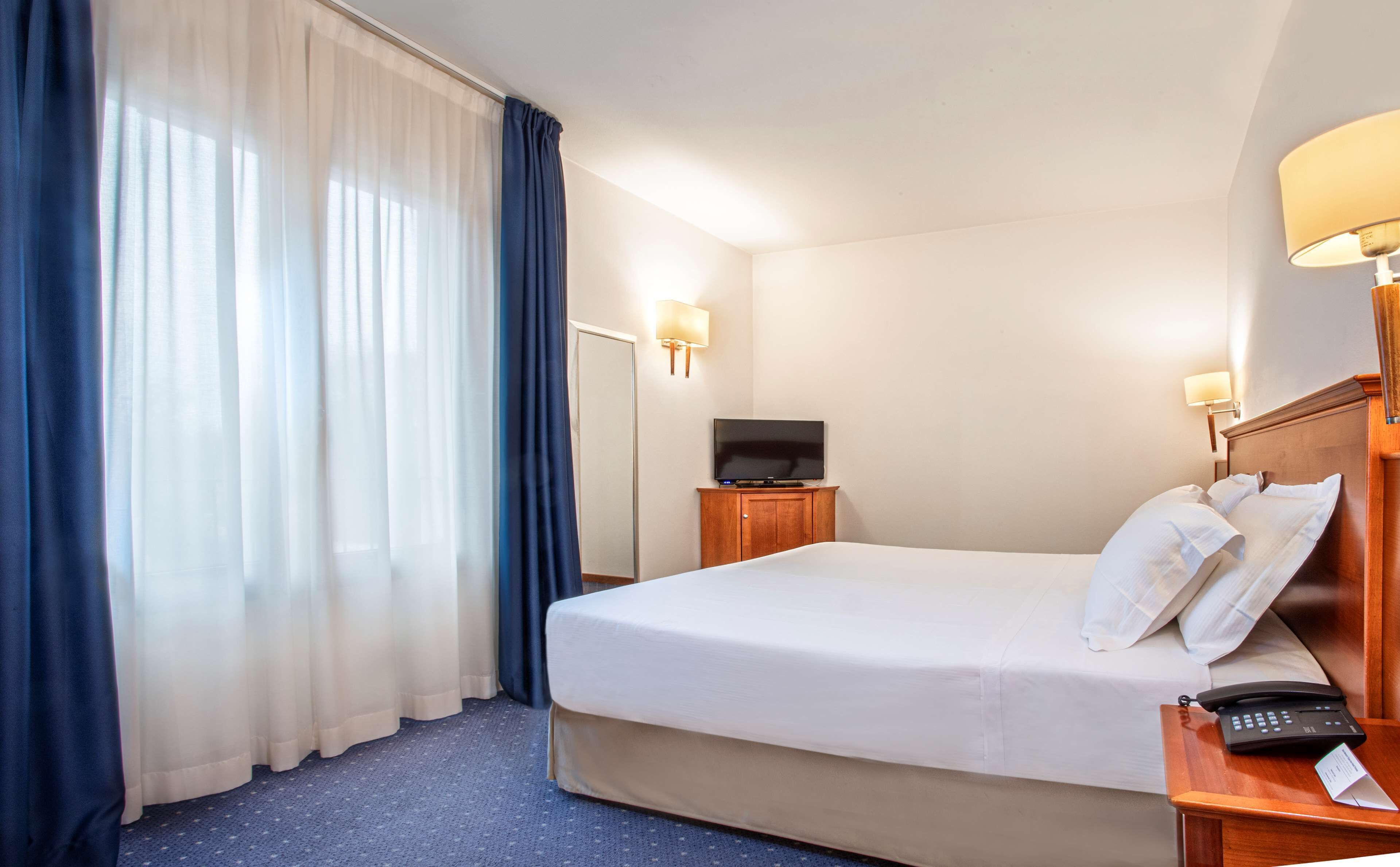 A hotel room with a large bed, a television on a wooden cabinet, a bedside table with a lamp, and a telephone.