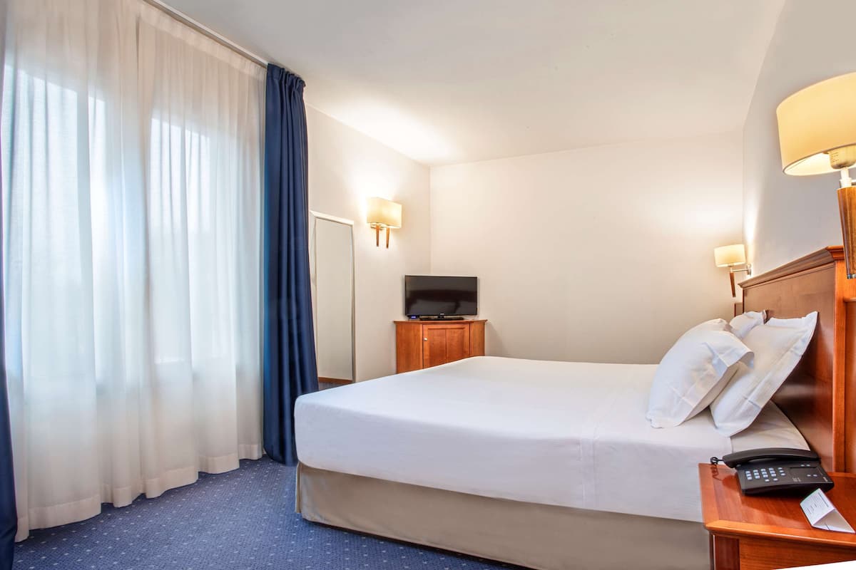 A hotel room with a large bed, a television on a wooden cabinet, a bedside table with a lamp, and a telephone.