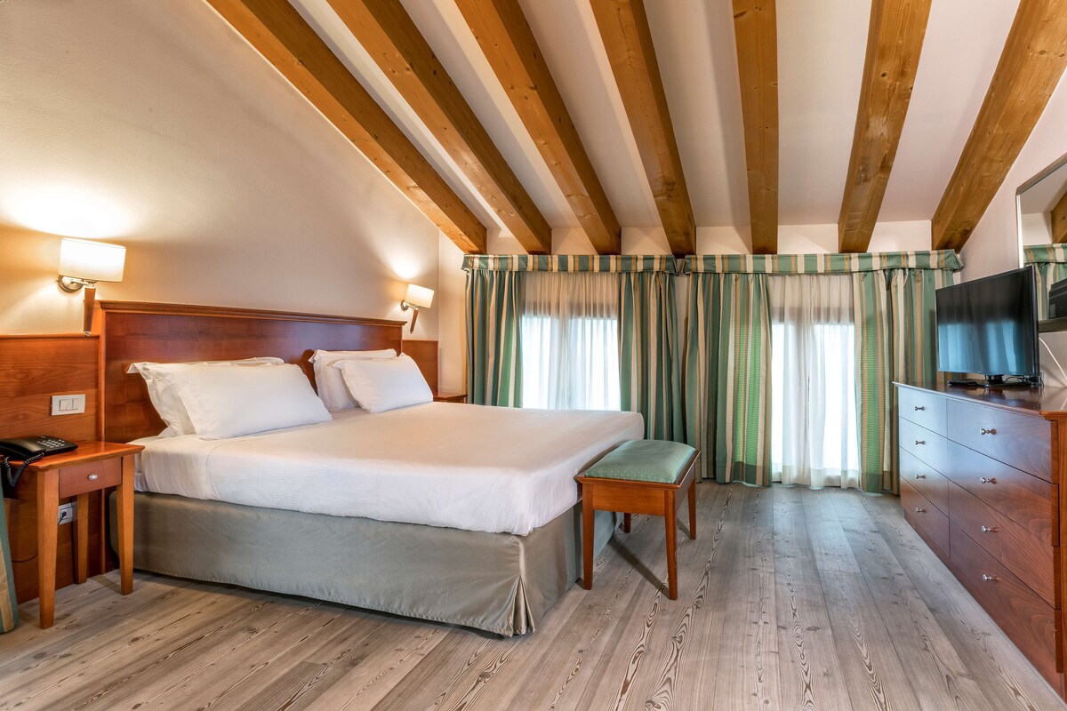 A hotel room with a wooden ceiling, a bed with white linens, a wooden nightstand, a desk with a telephone, a television on a wooden dresser, and a window with striped curtains.