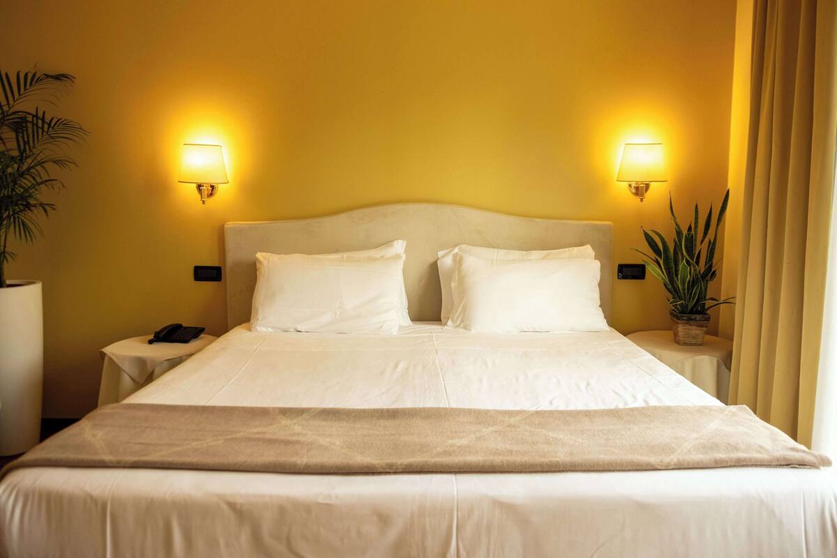 Habitación de hotel con una cama grande, dos lámparas de noche, una mesita con un teléfono y una planta en maceta.