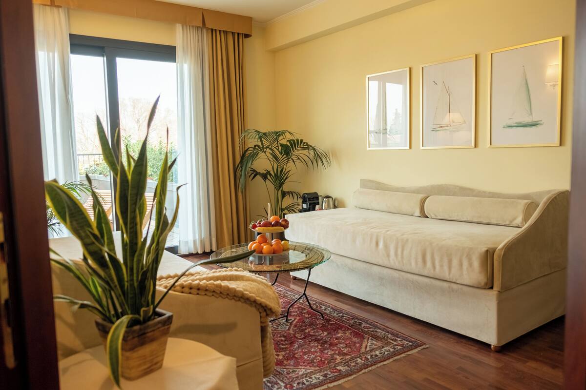 Una sala de estar con un sofá beige, una mesa de centro de vidrio con fruta, una planta en maceta y una alfombra roja.