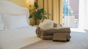 Un lit bien fait, avec du linge de lit blanc, une serviette pliée et une « boîte de confort » posée sur le sol d’une chambre d’hôtel.