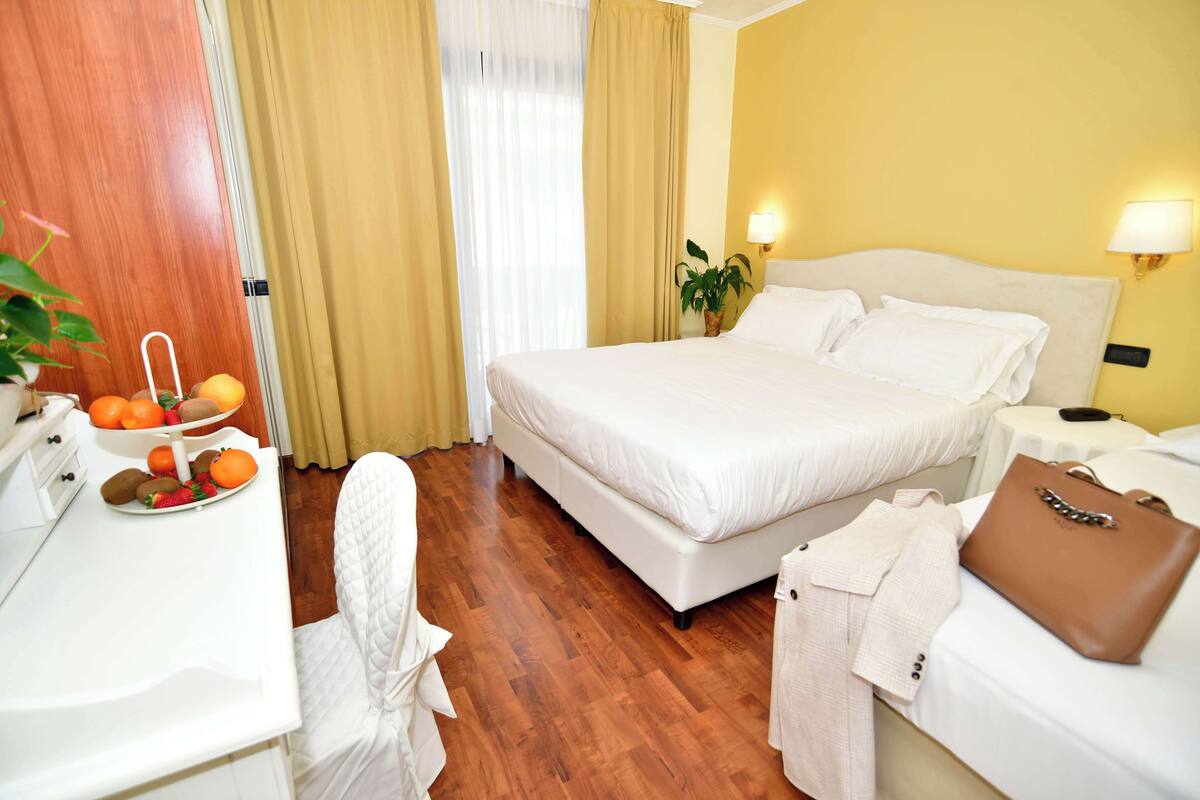Una habitación de hotel con cama, mesitas de noche, un escritorio con fruta y una planta.