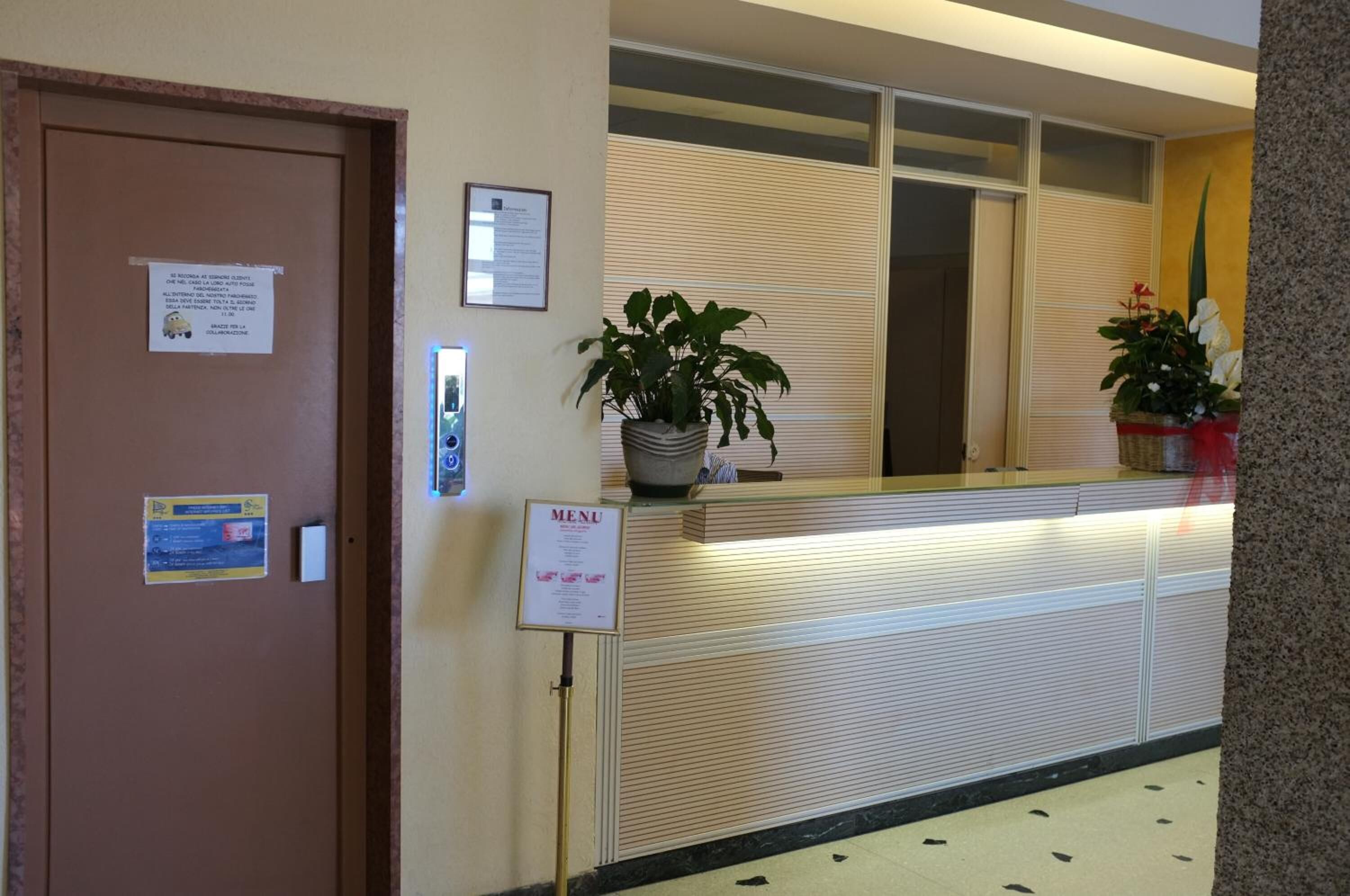 Un'area reception con un bancone, una porta con un cartello, un espositore per menu e piante in vaso.
