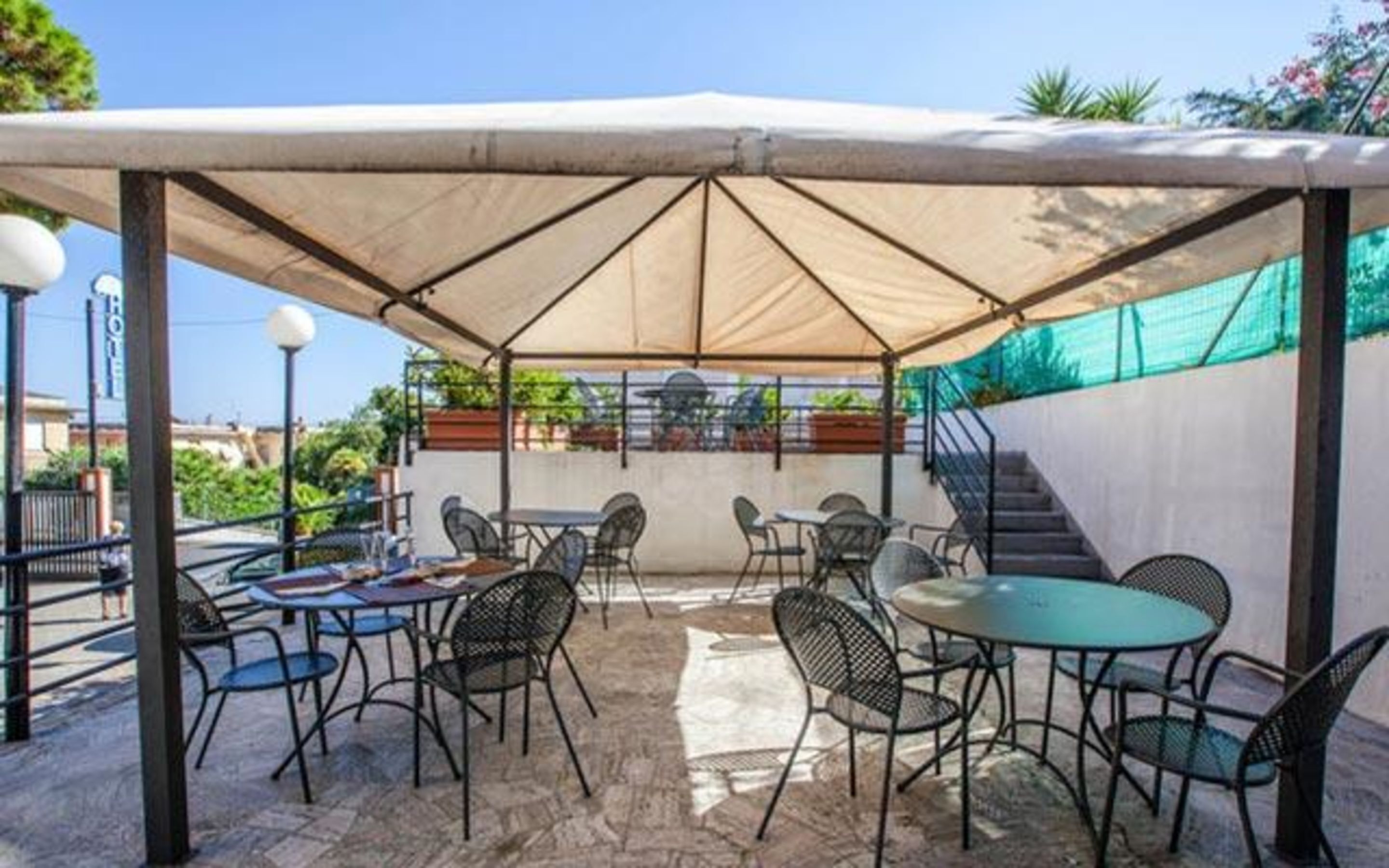 An outdoor patio area with tables and chairs under a large canopy, surrounded by a low wall and a staircase.