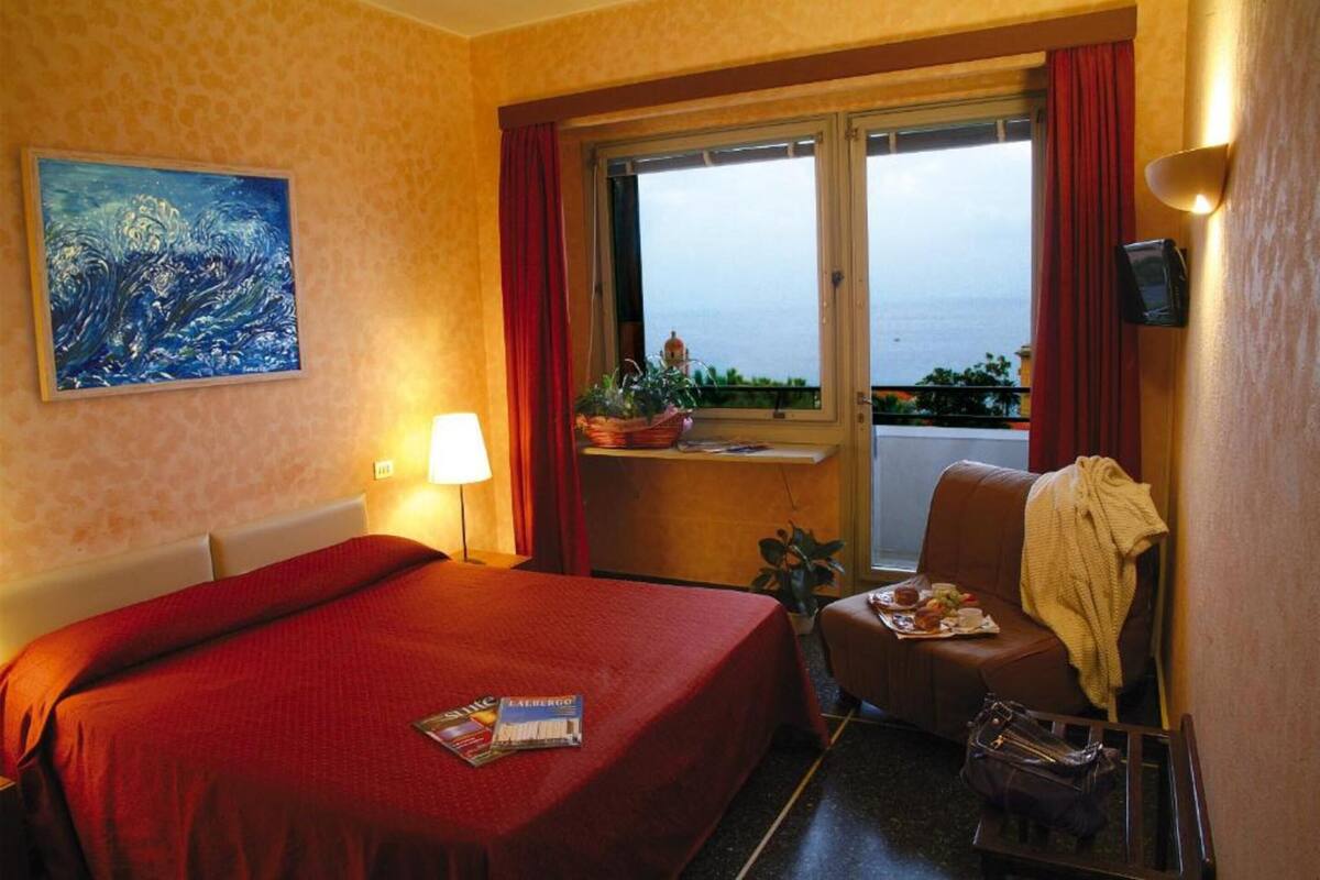 A hotel room with a large bed, a window with a view of the sea, a painting on the wall, and a small table with a plant and a tray of food.