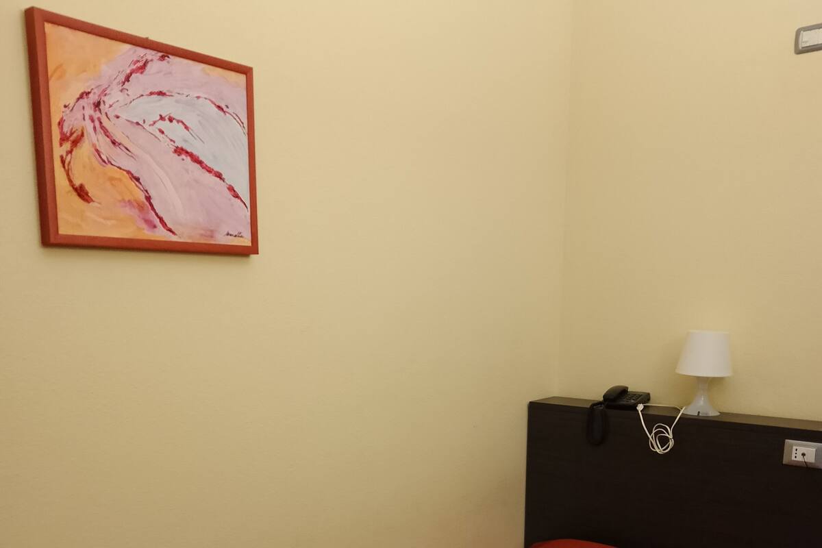 A hotel room with a single bed, a red bedspread, a rolled white towel, a painting on the wall, a black nightstand with a lamp, and a phone.