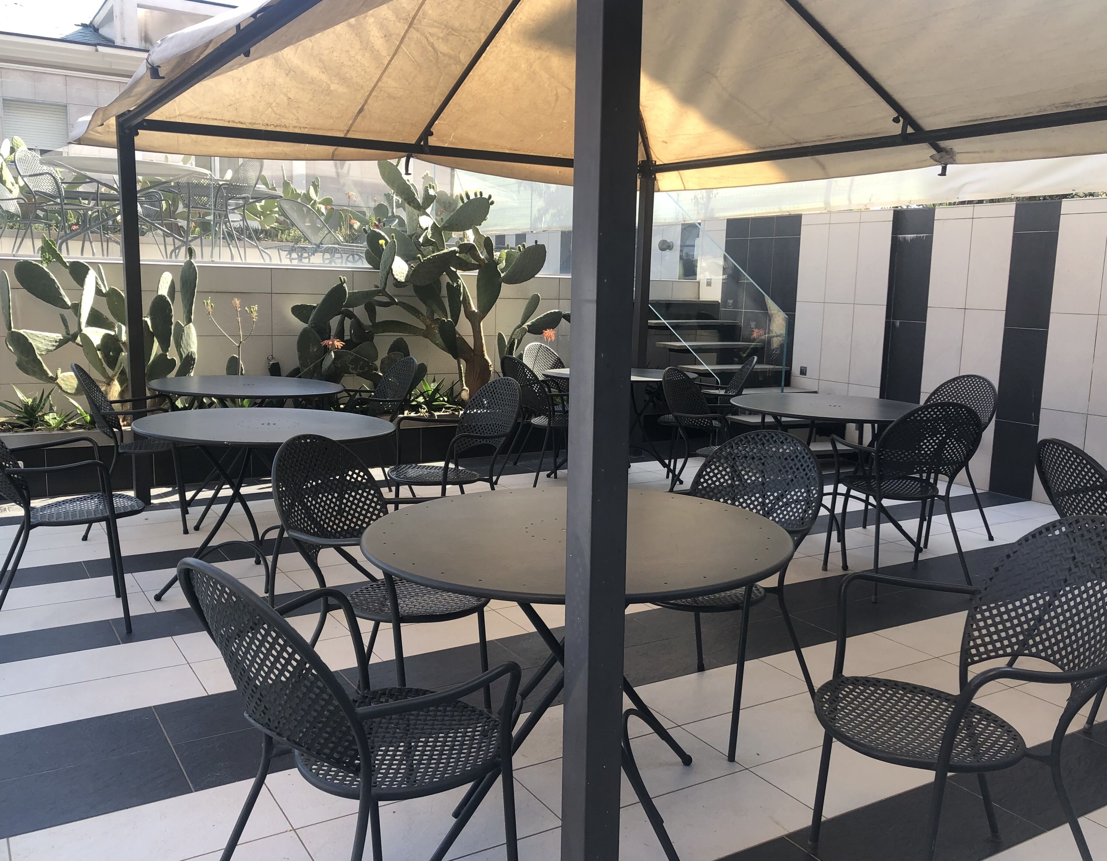 An outdoor patio area with round tables and black metal chairs, shaded by a canopy.