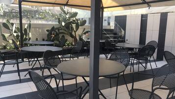 An outdoor patio area with round tables and black metal chairs, shaded by a canopy.