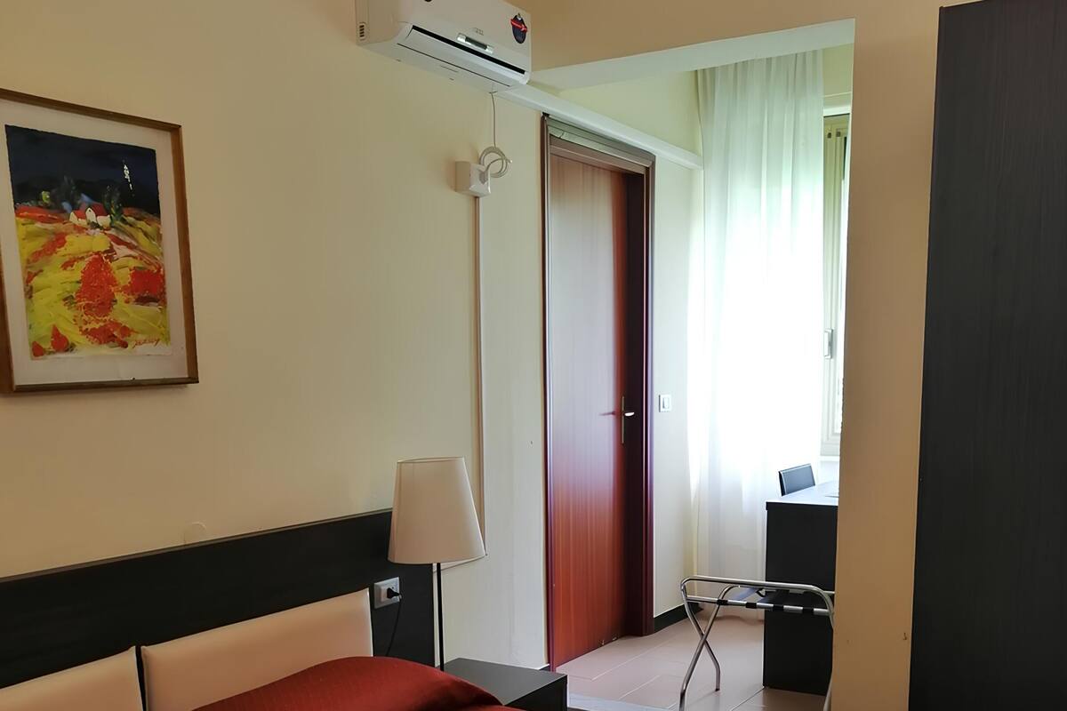 A hotel room with a large bed, a painting on the wall, an air conditioning unit, a desk with a chair, and a door leading to another room.
