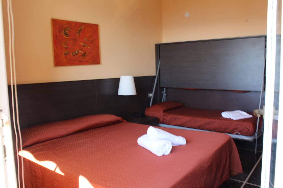 A hotel room with two beds, a red bedspread, a white towel on one bed, a red painting on the wall, and a dark headboard.