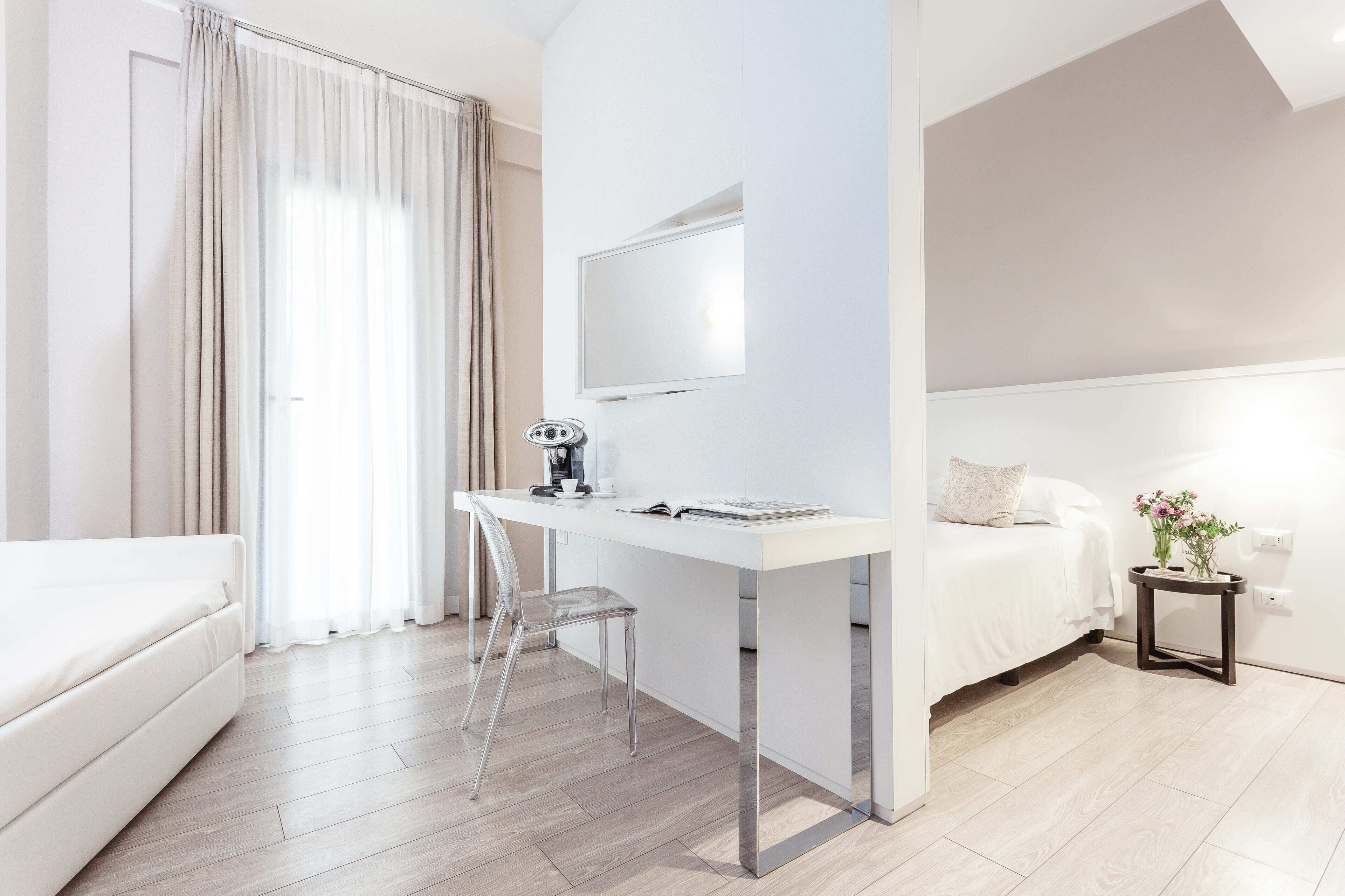 A modern hotel room with a white desk, a chair, a bed, and a small table with a vase of flowers.