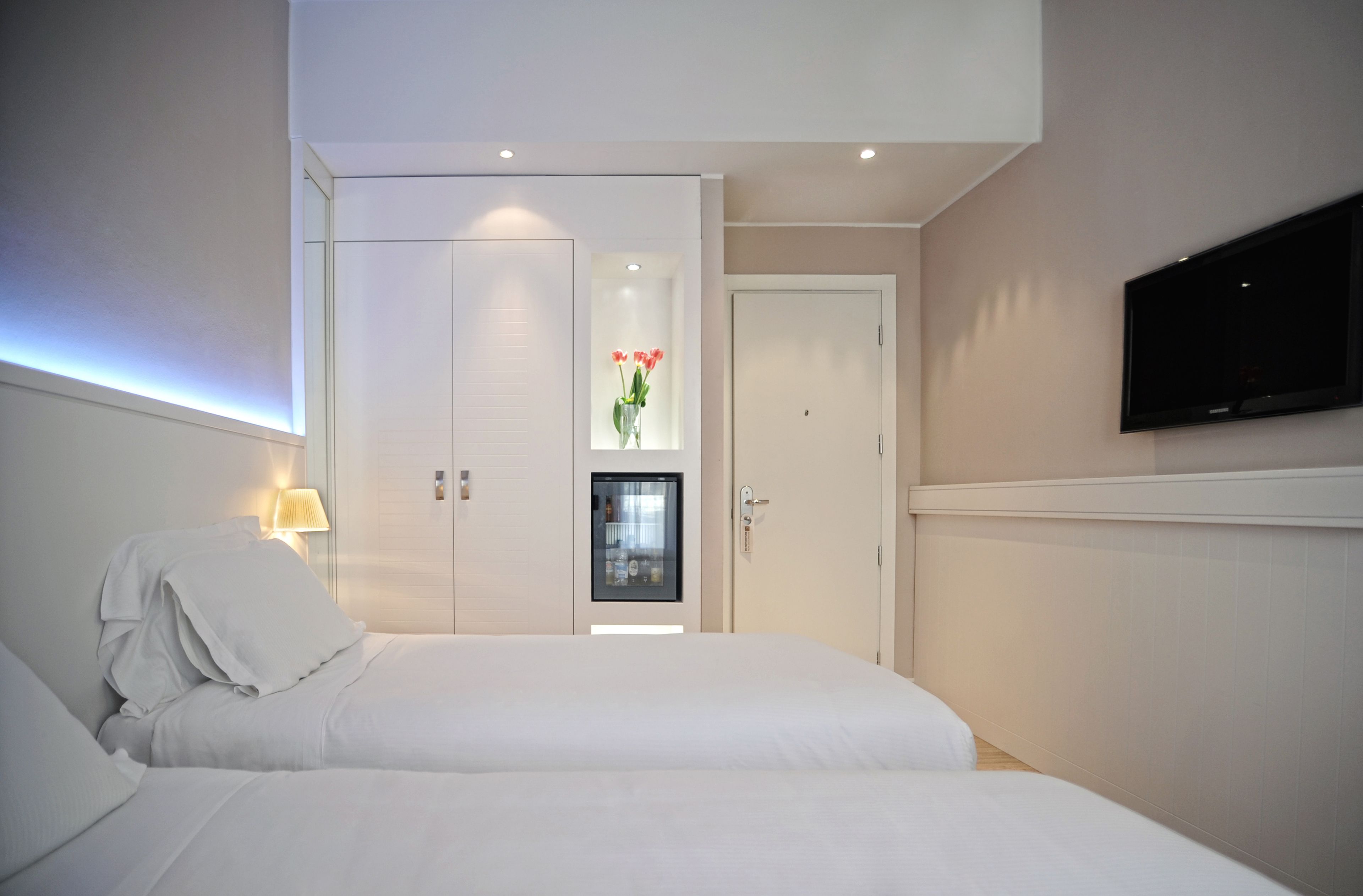 A hotel room with two beds, a TV mounted on the wall, a minibar, and a door with a frosted glass panel.